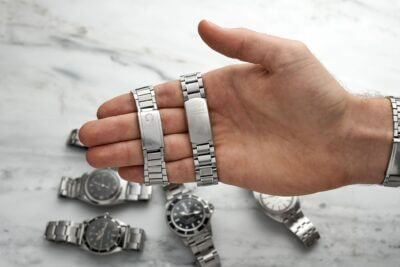 A hand holds two classic silver watch bracelets, surrounded by various watches on a marble surface, showcasing the timeless appeal of watch accessories