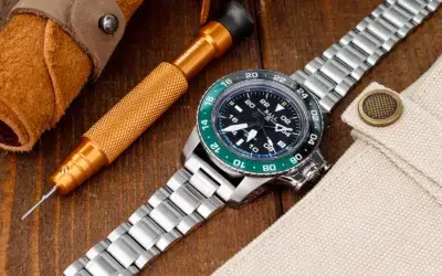 A close-up of a metal watch with a black dial and green bezel, placed on a wooden surface alongside a tool and fabric, highlighting watch styling and accessories