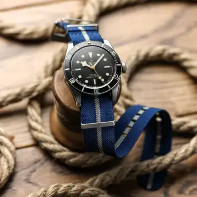 A stylish watch with a black dial and silver accents, featuring a blue nylon strap, rests on a wooden surface surrounded by nautical rope