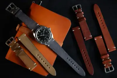 A stylish watch with a black face is placed on a brown leather notebook, surrounded by various vintage leather watch straps in shades of brown and tan