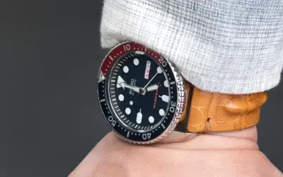 A close-up of a wrist wearing a watch with a black dial and red accents, paired with a stylish crocodile leather strap. The background is softly blurred to highlight the watch
