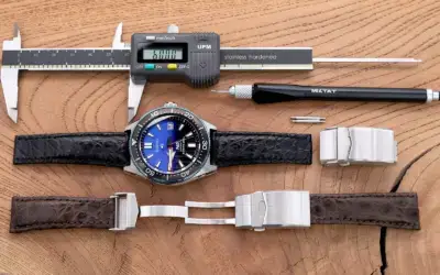 A close-up of a watch with a blue dial, surrounded by tools and two watch straps. A caliper measures the strap's width, showcasing a deployant clasp installation process