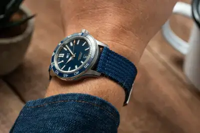 A close-up of a wrist wearing a blue nylon watch strap, showcasing a stylish watch with a blue dial. The background features a plant and a coffee cup on a wooden table