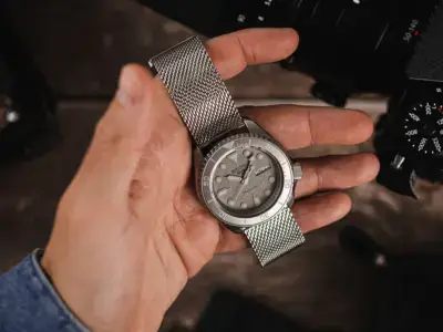 A hand holding a silver mesh quick release watch band with a light gray watch face, set against a blurred background of camera equipment