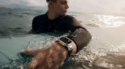 A person in a wetsuit reaches for a surfboard in the ocean, showcasing a stylish watch on their wrist, ideal for beach vacations and water activities