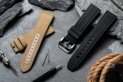 A collection of tech-savvy watch straps and accessories, featuring a tan fabric strap and a black rubber strap, displayed on a textured stone background with tools