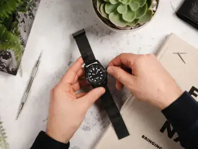 A person adjusts a quick release watch strap on a black watch, surrounded by a succulent plant, a notebook, and a pen on a marble surface