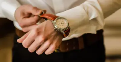 A close-up of a person adjusting a stylish watch with a brown leather strap on their wrist, dressed in a white shirt and dark pants, showcasing a fashionable accessory