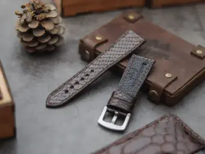 A close-up of a snake skin watch strap placed on a textured surface, alongside a wooden box and a pinecone, highlighting its unique design and craftsmanship