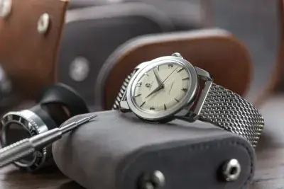 A stylish watch with a mesh strap rests on a leather pouch, surrounded by watchmaking tools and leather straps, highlighting modern fashion trends in timepieces