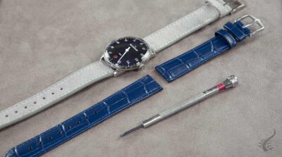 A watch with a black dial and silver case, alongside two interchangeable straps: a light gray leather strap and a blue crocodile-pattern strap, with a tool for changing straps