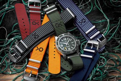 A collection of colorful nylon watch bands in various patterns, arranged around a classic metal watch, set against a backdrop of tangled green fishing nets