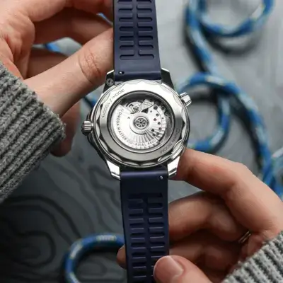 A person holds a waterproof watch with a blue strap, showcasing its intricate back design against a textured background featuring coiled ropes