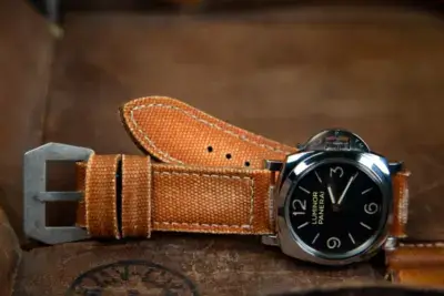 A vintage watch with a black dial and silver casing rests on a brown leather surface, showcasing a worn orange strap that adds a nostalgic touch