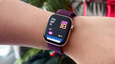 A close-up of a wrist wearing a stylish smartwatch with a purple strap, displaying the date and time, set against a backdrop of greenery and festive decorations