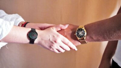 Two hands reaching out to each other, one wearing a black smartwatch and the other a gold watch, against a neutral background
