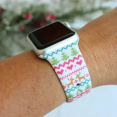 A close-up of a festive watch band featuring a colorful holiday pattern with reindeer, hearts, and trees, worn on a wrist against a blurred holiday background