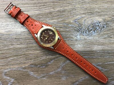 A close-up of a stylish cuff watch strap in orange leather with perforations, featuring a round brown watch face, set against a wooden background