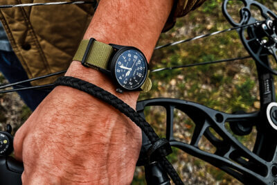 A close-up of a hand wearing a black outdoor watch with a green strap, gripping a bowstring, showcasing the watch's durability for outdoor adventures