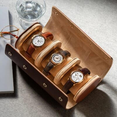 A stylish leather watch roll displaying four elegant watches, arranged neatly on a soft interior, with a glass of water and a book in the background