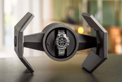A sleek, modern watch winder displaying a luxury watch. The design features angular shapes, highlighting the watch's intricate details against a blurred background