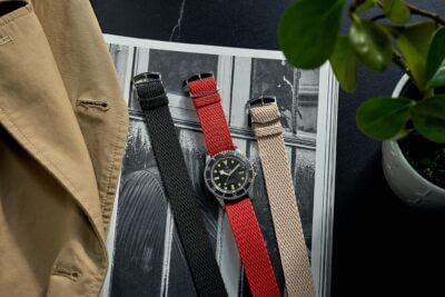 A stylish watch with interchangeable bands in black, red, and beige, placed on a magazine with a plant and a beige jacket nearby, showcasing everyday elegance
