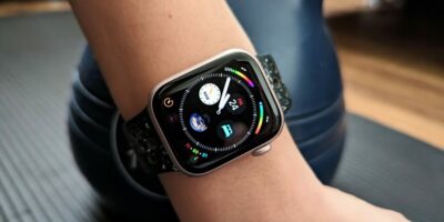 A close-up of an Apple Watch on a wrist, displaying a colorful watch face with various app icons, set against a blurred background of fitness equipment