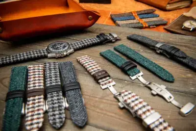 A variety of watch straps displayed on a wooden surface, featuring heavy-duty and vintage styles in different colors and patterns, alongside a classic watch and leather accessories