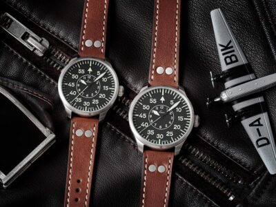 A close-up of two pilot watches with black dials and brown leather straps, placed on a black leather surface alongside a model airplane and a silver buckle