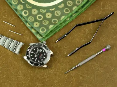 A watch, spring bars, tweezers, and a screwdriver are arranged on a textured surface, highlighting essential tools for watch maintenance and care