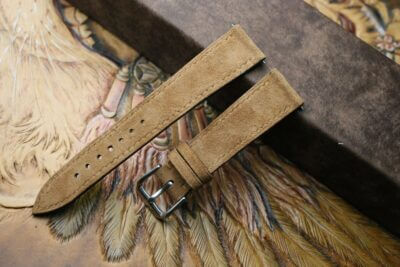 A close-up of a tan suede watch strap with a silver buckle, resting on an ornate background, highlighting the craftsmanship and texture of the material
