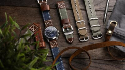 A collection of canvas watch straps in various colors and styles laid out on a wooden surface, alongside a watch and some accessories, highlighting their historical appeal