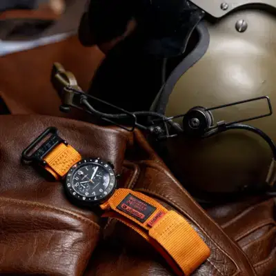 A rugged smartwatch with an orange heavy-duty strap placed on a brown leather surface, alongside a vintage helmet, showcasing durability and style