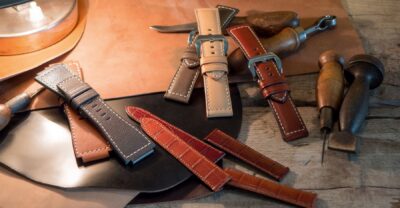 A collection of leather watch straps in various colors and styles, arranged on a wooden surface with tools and materials for watchmaking in the background