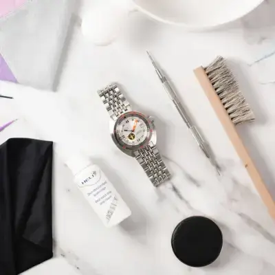 A silver metal watch with a detailed face, surrounded by cleaning tools, a bottle of watch care solution, and cloths on a marble surface, illustrating watch maintenance