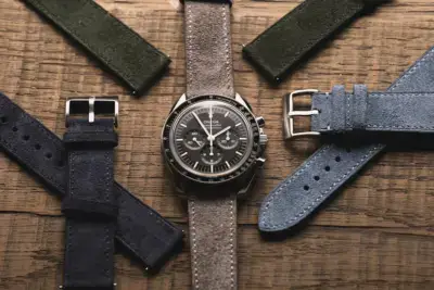 A stylish watch with a dark dial is surrounded by various suede watch straps in green, blue, and navy, all set against a rustic wooden background