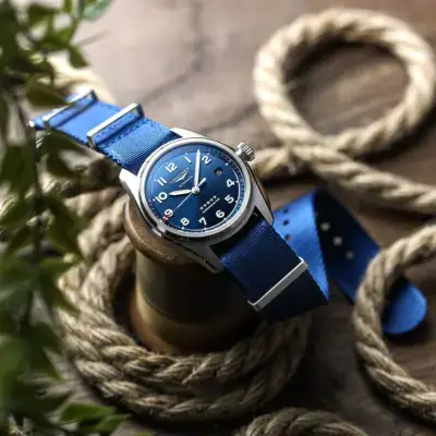 A close-up of a blue nylon watch strap attached to a silver watch, resting on a wooden surface surrounded by coiled rope and greenery