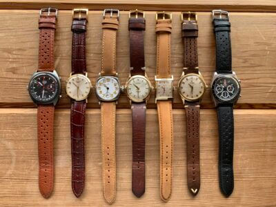 A collection of seven watches with various leather straps, displayed on a wooden surface, showcasing different styles and colors for daily wear and tear protection