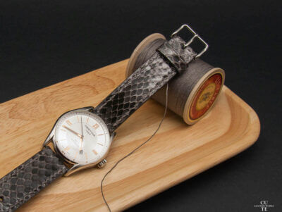 A snake skin watch strap placed on a wooden tray next to a spool of thread and needle, showcasing the elegance and care needed for maintenance