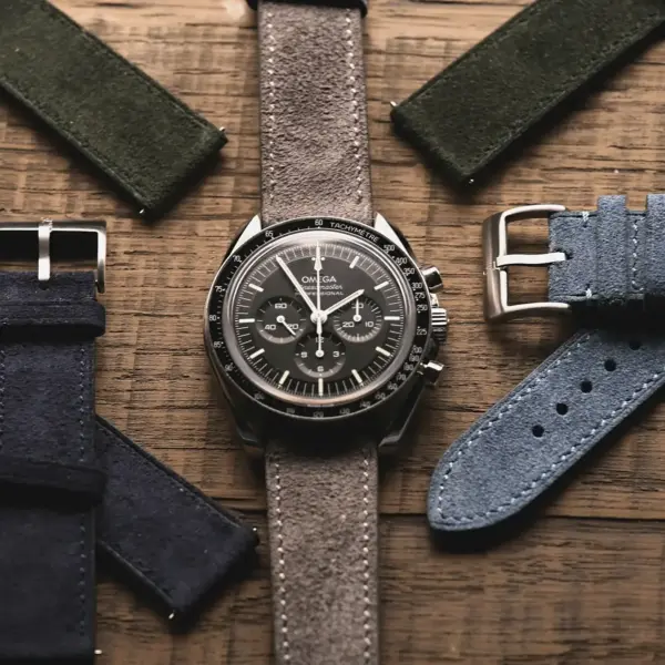 A stylish watch with a dark dial is surrounded by various suede watch straps in green, blue, and navy, all set against a rustic wooden background