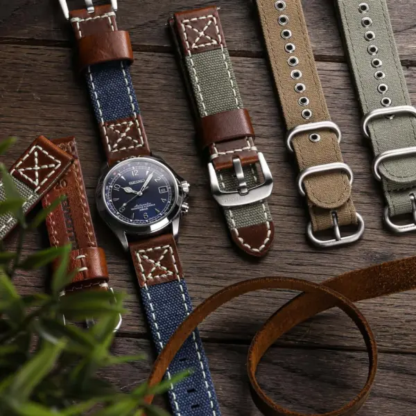 A collection of canvas watch straps in various colors and styles, displayed alongside a watch, a pen, and a plant on a wooden surface