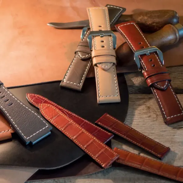 A collection of leather watch straps in various colors and styles, arranged on a wooden surface with tools and materials for watchmaking in the background