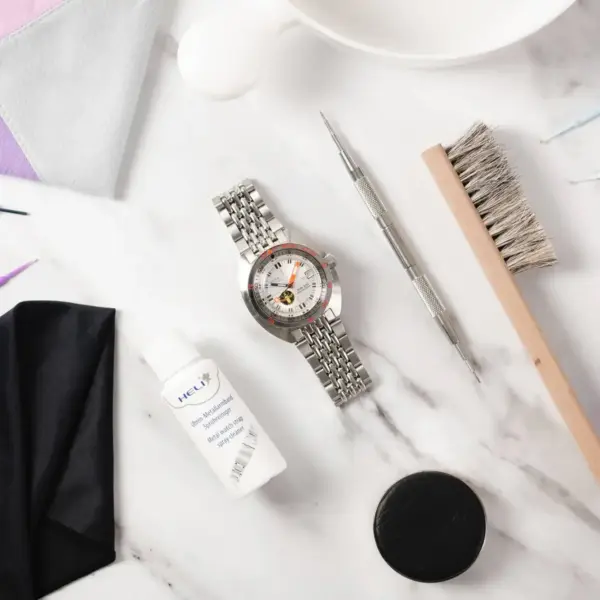 A silver metal watch with a detailed face, surrounded by cleaning tools, a bottle of watch care solution, and cloths on a marble surface, illustrating watch maintenance