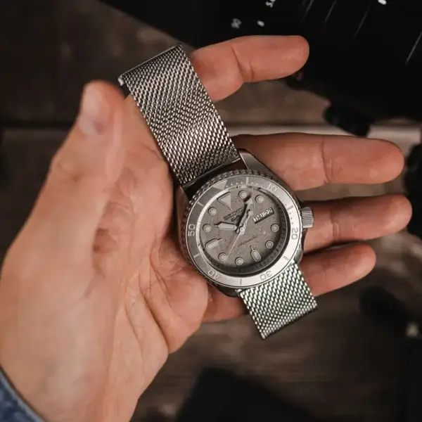 A hand holding a silver mesh quick release watch band with a light gray watch face, set against a blurred background of camera equipment
