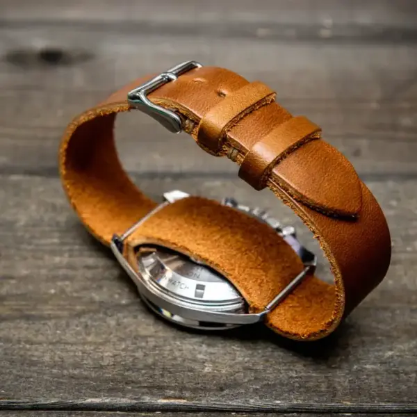 A close-up of a one-piece leather watch strap featuring a silver watch, resting on a wooden surface, showcasing its texture and craftsmanship