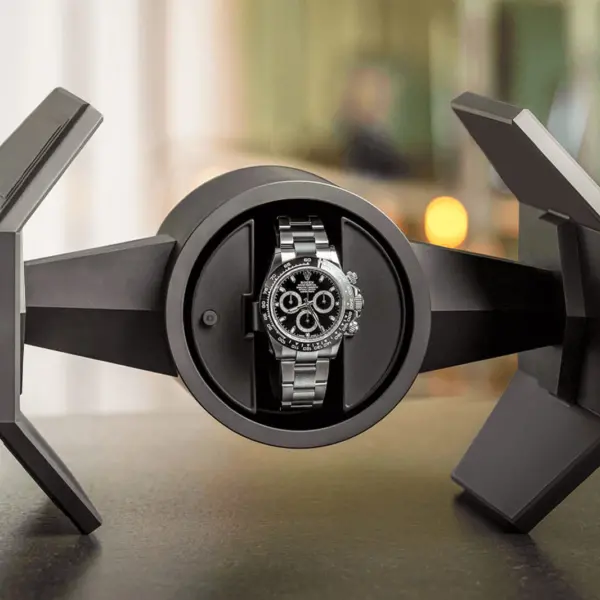 A sleek, modern watch winder displaying a luxury watch. The design features angular shapes, highlighting the watch's intricate details against a blurred background