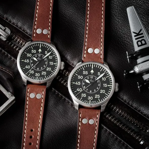 A close-up of two pilot watches with black dials and brown leather straps, placed on a black leather surface alongside a model airplane and a silver buckle
