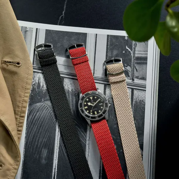 A stylish watch with interchangeable bands in black, red, and beige, placed on a magazine with a plant and a beige jacket nearby, showcasing everyday elegance