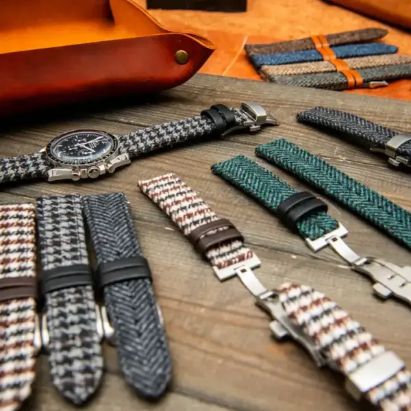 A variety of watch straps displayed on a wooden surface, featuring heavy-duty and vintage styles in different colors and patterns, alongside a classic watch and leather accessories