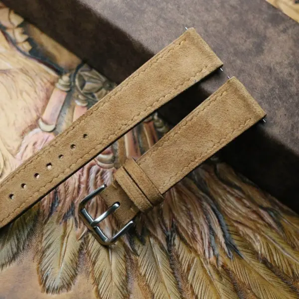 A close-up of a tan suede watch strap with a silver buckle, resting on an ornate background, highlighting the craftsmanship and texture of the material